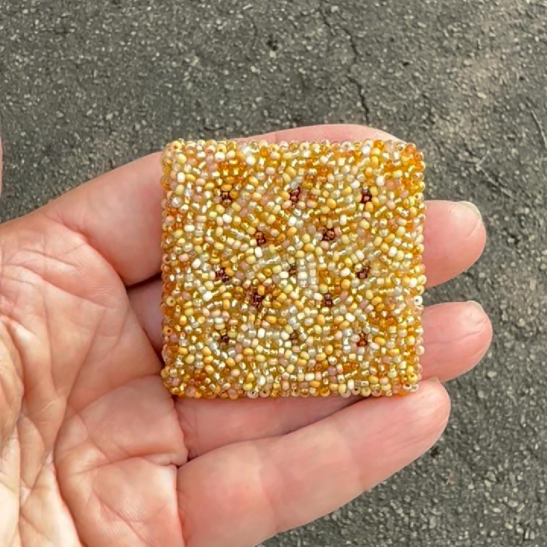 A hand holding a small square cracker brooch with seed beads in a warm yellow-gold and white colorway, with gray asphalt in the background.