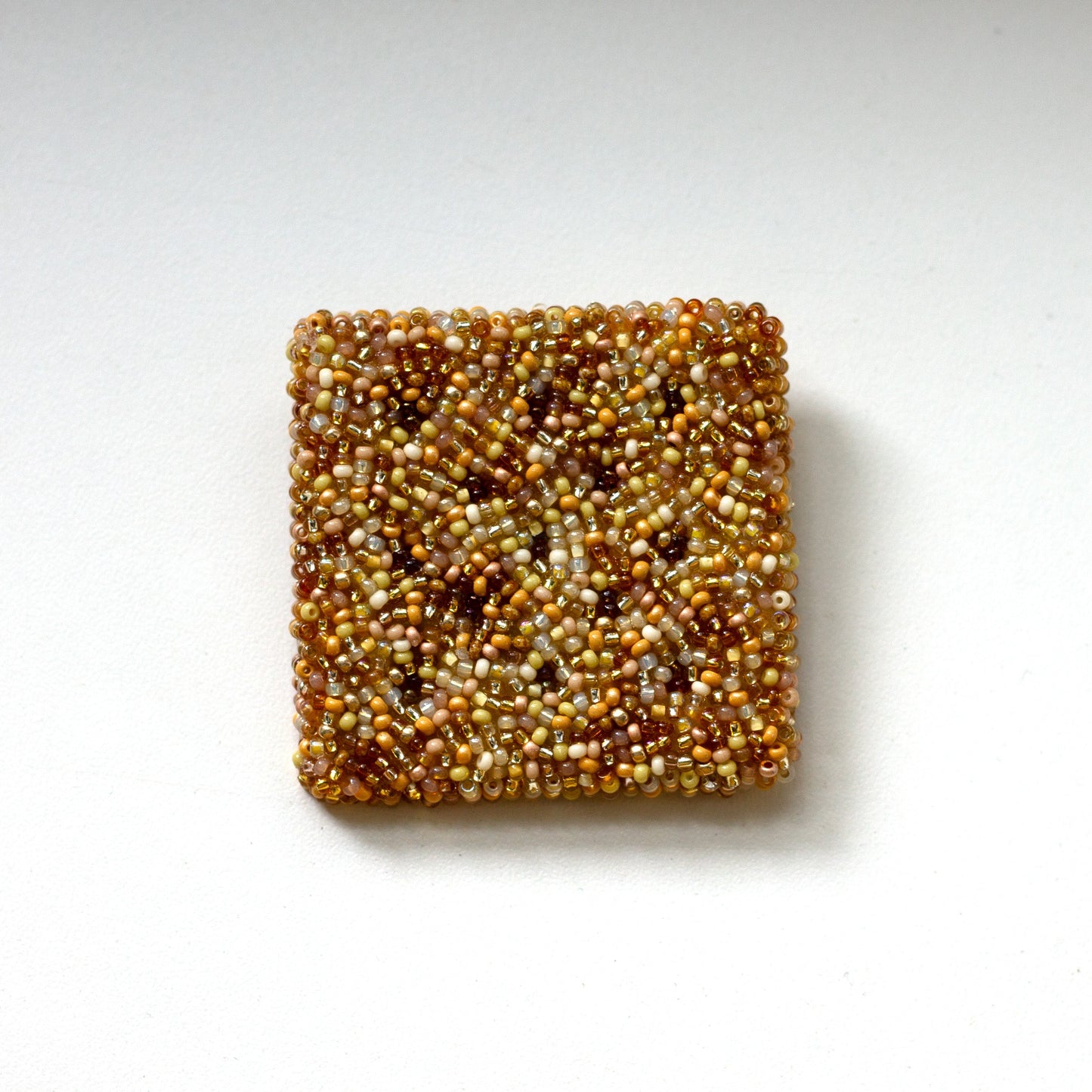 A square beaded cracker brooch made of tightly packed seed beads in a mottled mix of gold, bronze, and brown tones, centered on a plain white background.