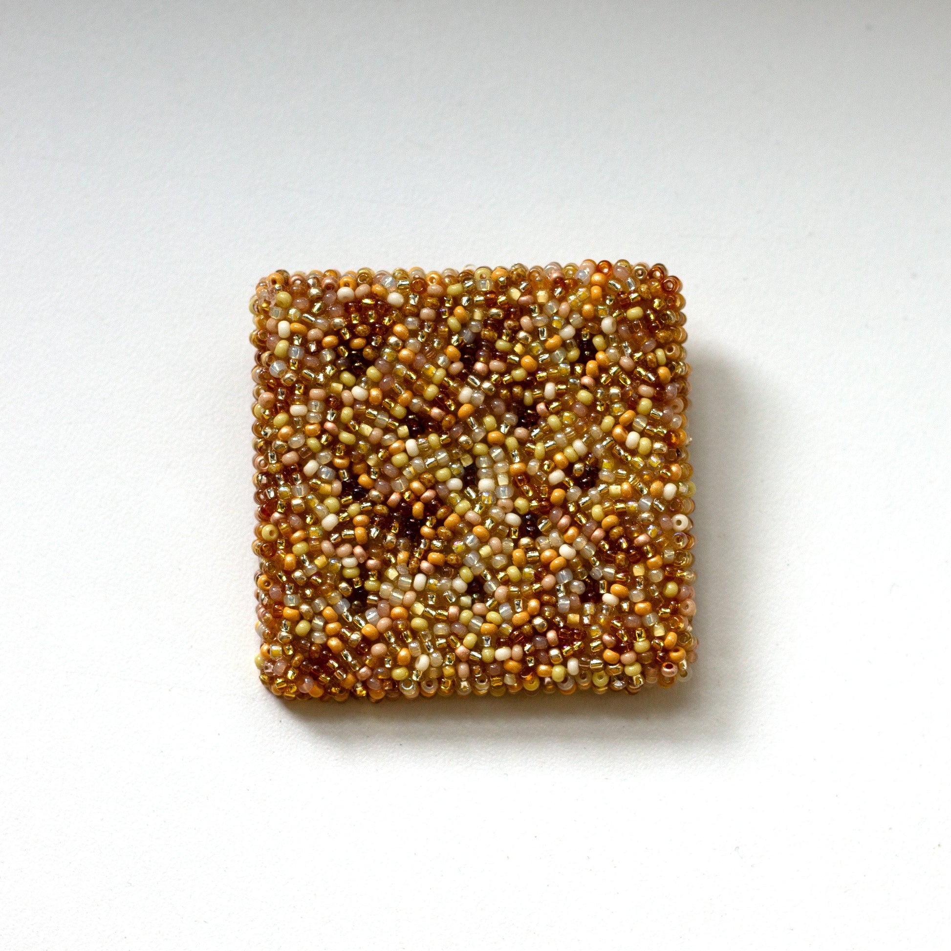 A square beaded cracker brooch made of tightly packed seed beads in a mottled mix of gold, bronze, and brown tones, centered on a plain white background.