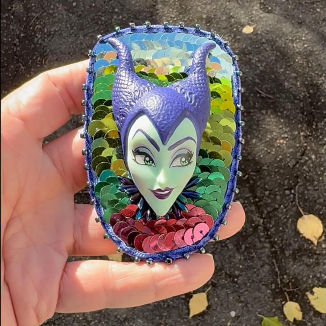 Hand holding a handmade purple and green sequined brooch featuring a Maleficent doll shown in the sunlight.
