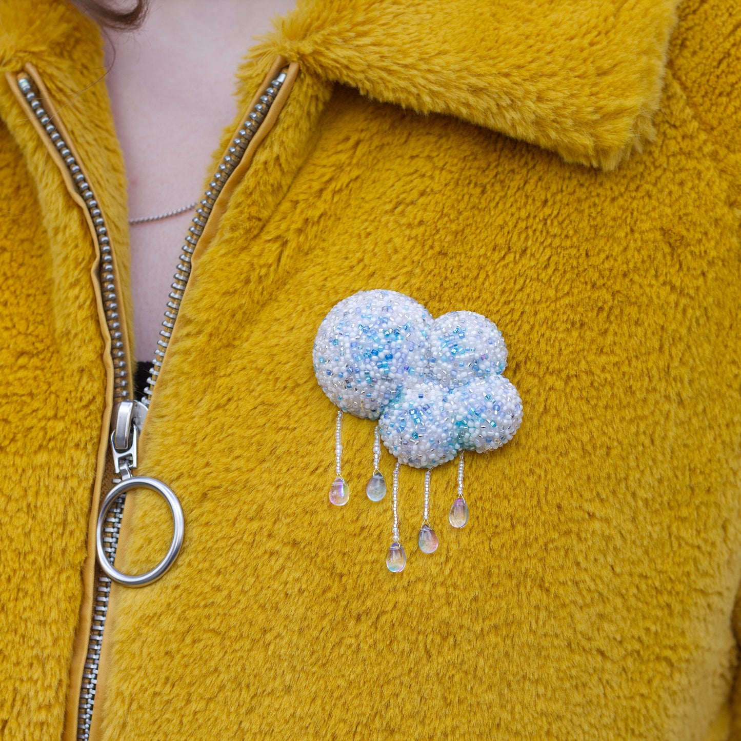 A close-up of a person wearing a fuzzy mustard yellow zip-up jacket with a light blue and white cloud-shaped beaded brooch pinned to the chest.