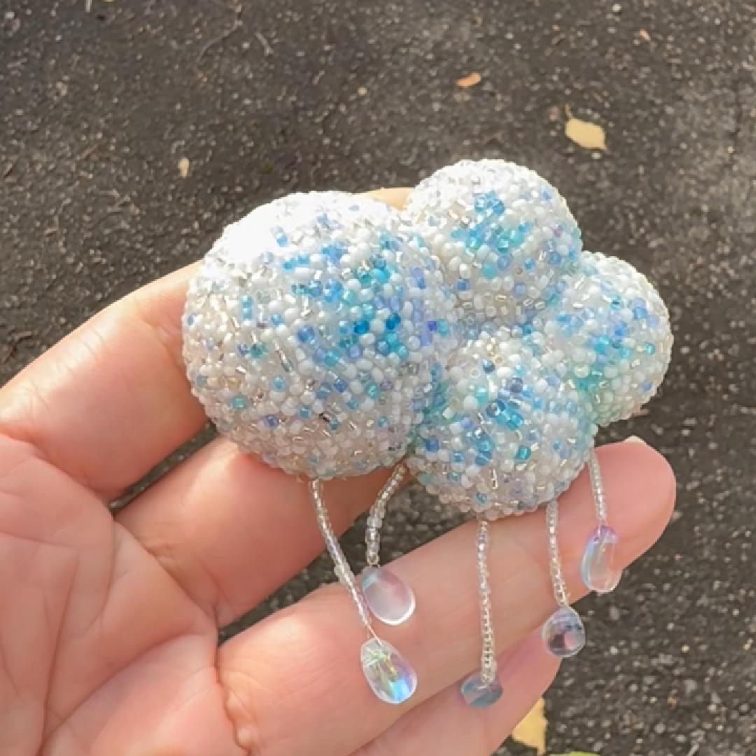 A handmade cloud-shaped beaded brooch held in a hand, featuring white and light blue beads with dangling iridescent bead drops, set against an outdoor asphalt background.