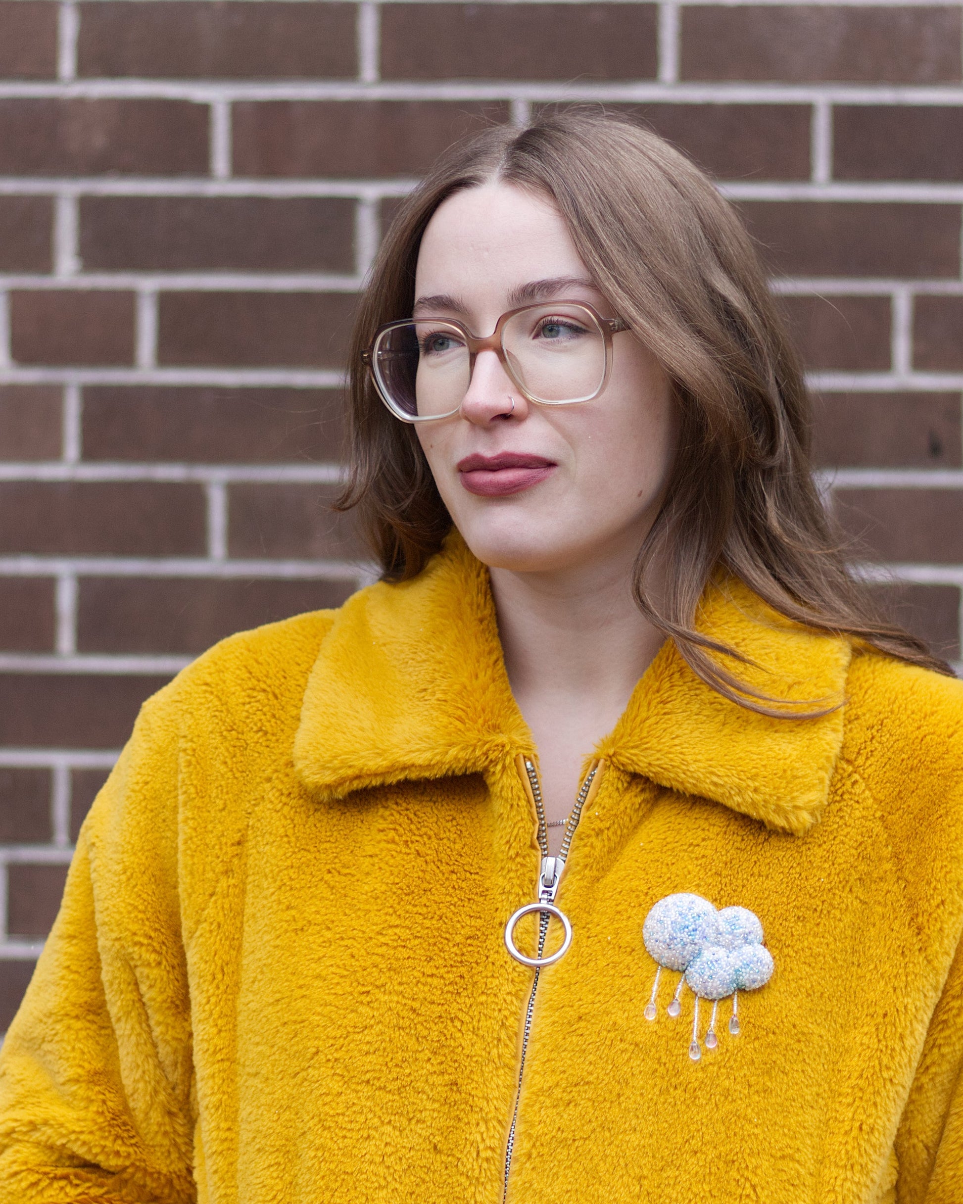 A woman with glasses wearing a fuzzy mustard yellow zip-up jacket with a beaded cloud brooch, standing in front of a brown brick wall.