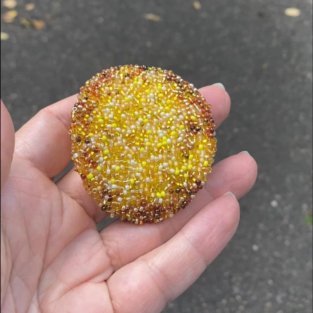 Hand holding a beaded potato chip brooch, with yellow and brown colors in the sunlight.