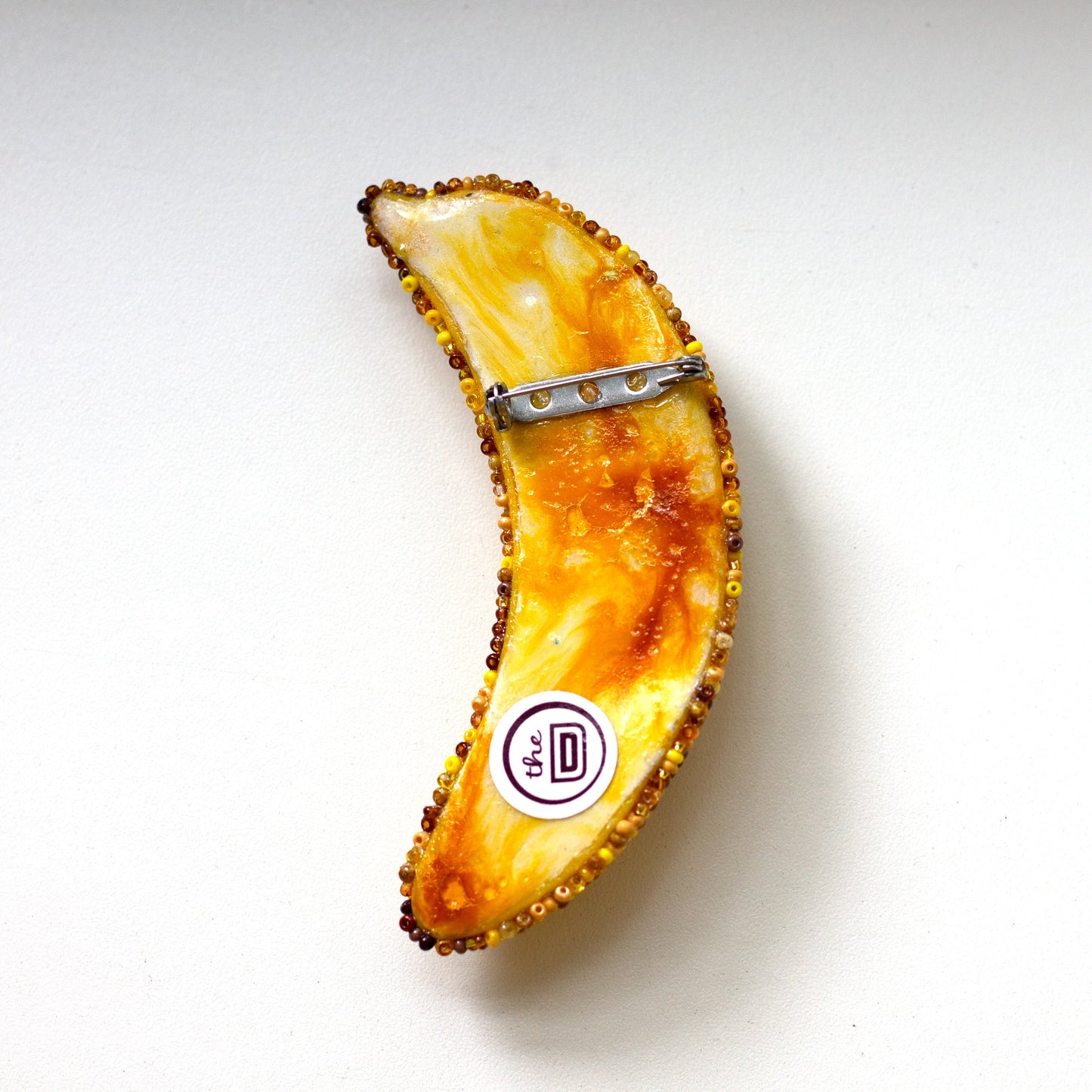 The back of a bruised banana pin featuring a marbled orange, yellow, and white pattern with a textured dark border and a silver metal clip, shown on a plain white background.