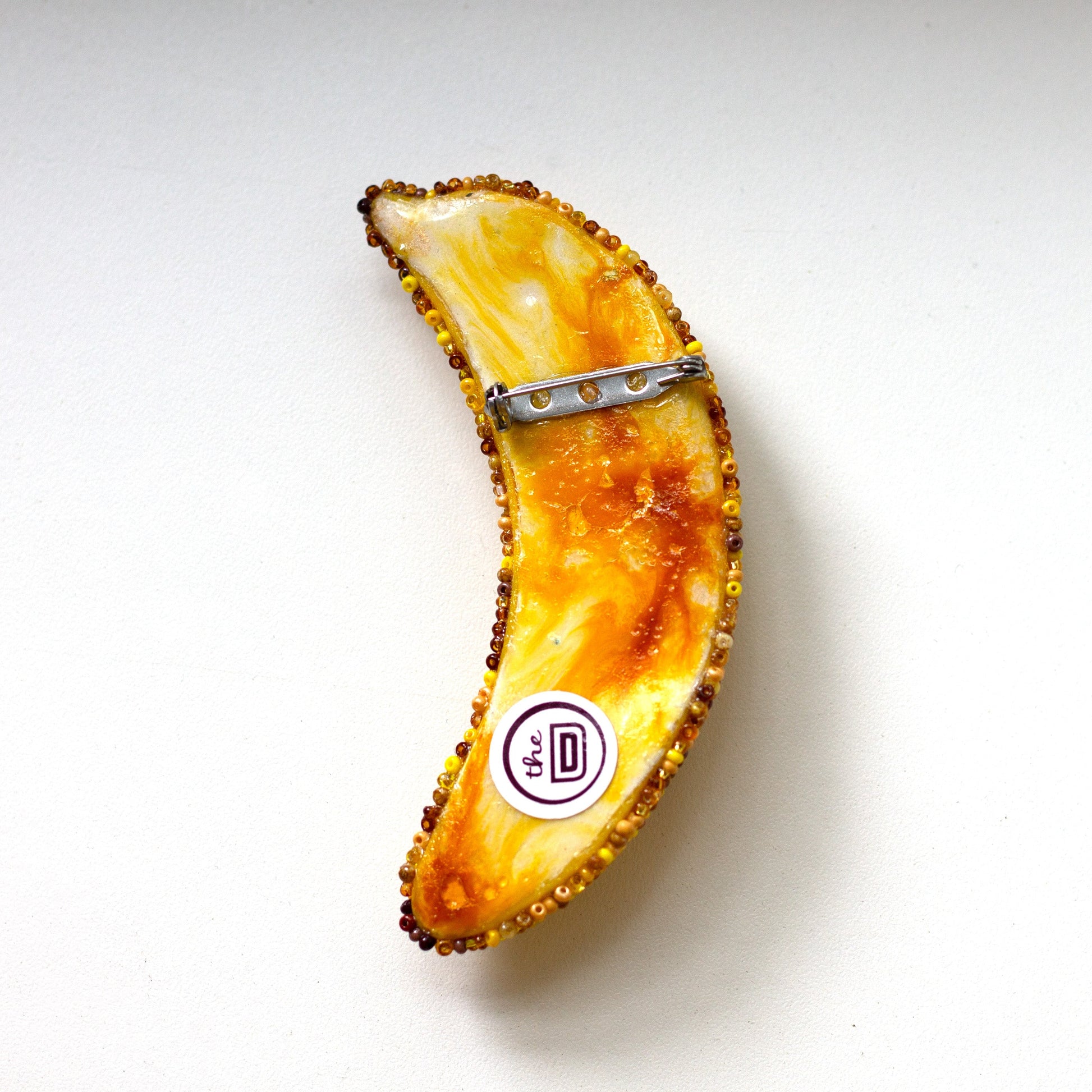 The back of a bruised banana pin featuring a marbled orange, yellow, and white pattern with a textured dark border and a silver metal clip, shown on a plain white background.