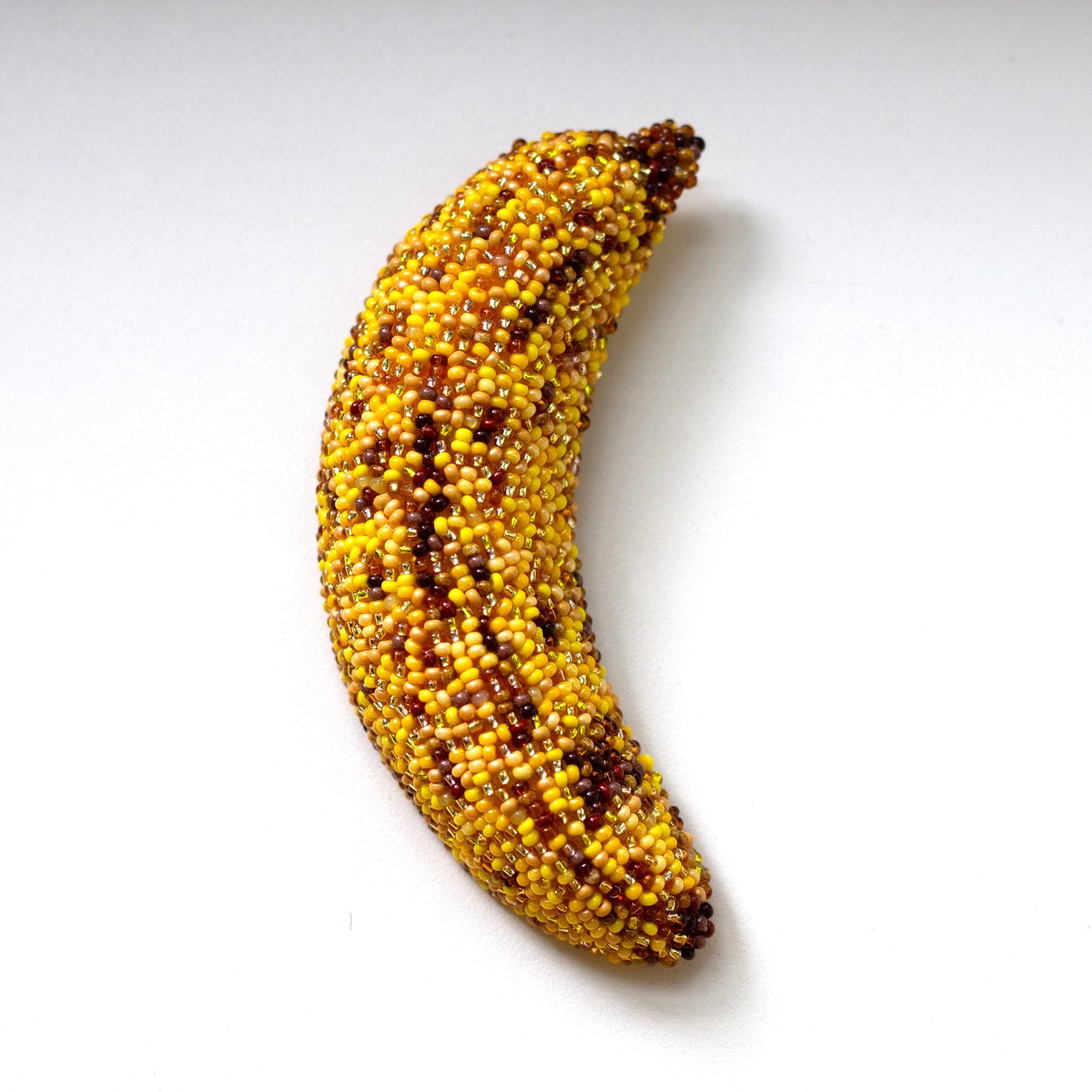 A bruised banana brooch covered in small yellow and brown seed-like beads, set against a plain white background.
