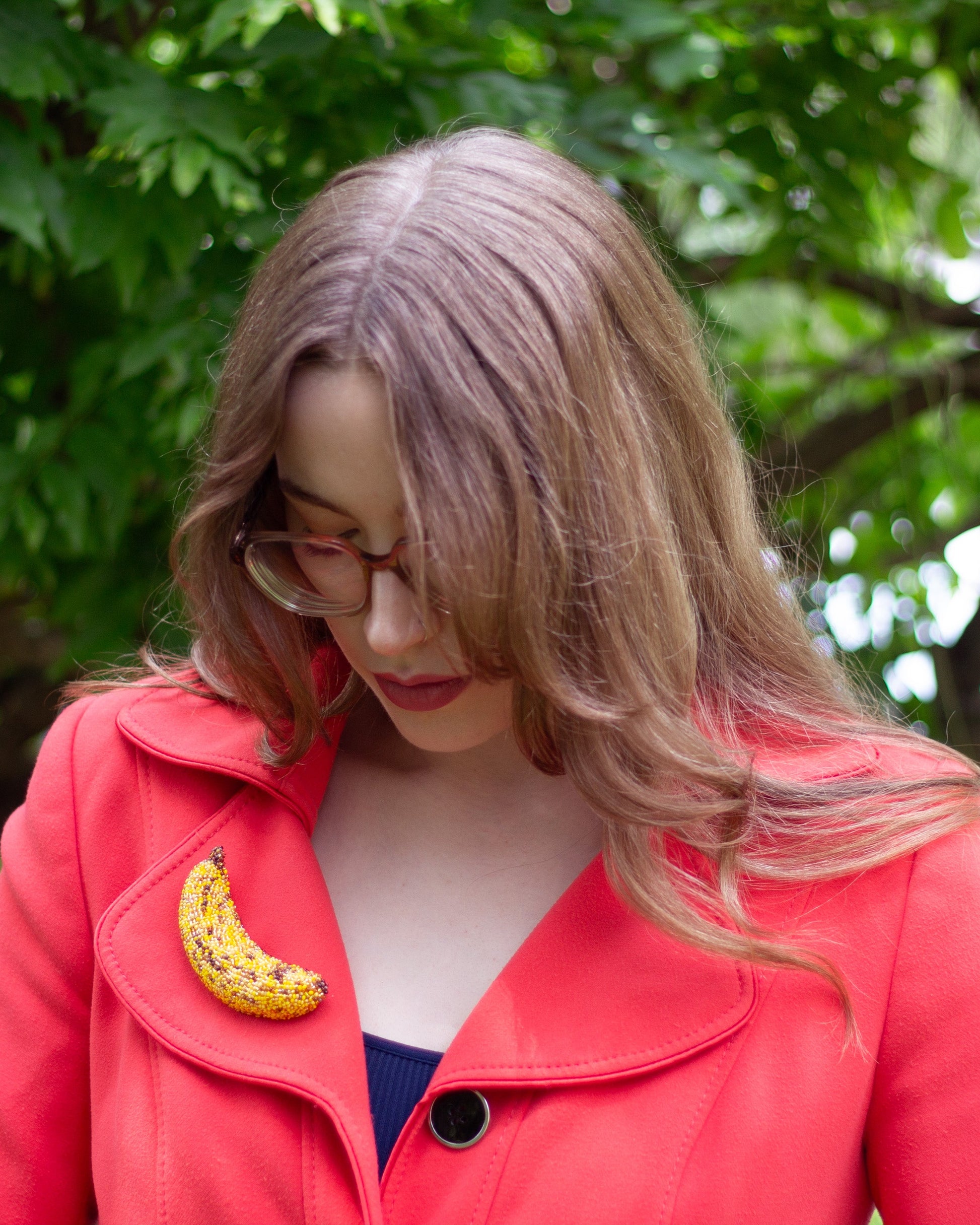 A woman with light brown hair and glasses, wearing a bright red coat over a dark top, wearing a yellow bruised banana brooch on her lapel, set against a background of green trees.