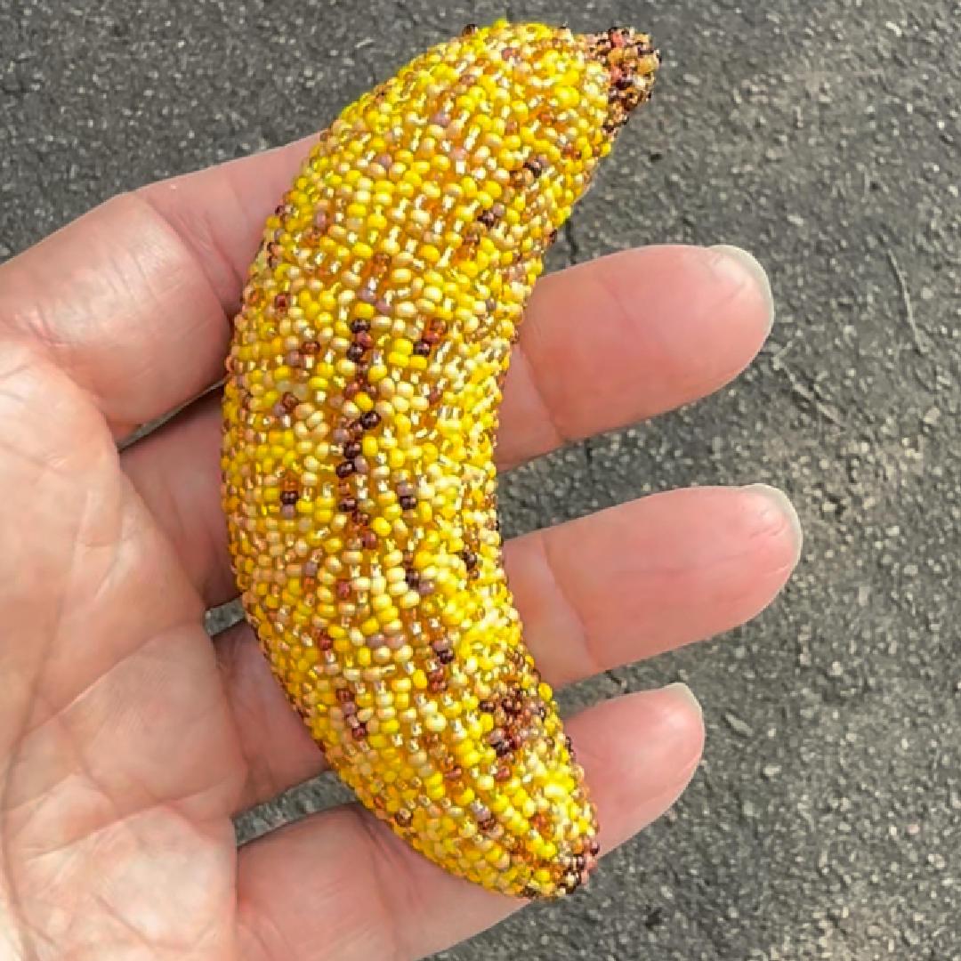 A hand holding a beaded bruised banana brooch outdoors on a gray asphalt surface.