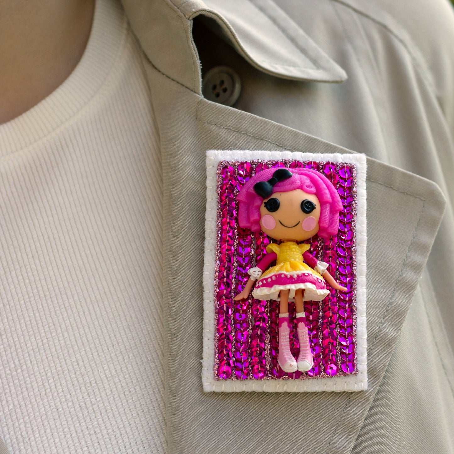 Closeup of a handmade Lalaloopsy doll brooch covered in pink sequins worn on a trench coat lapel.