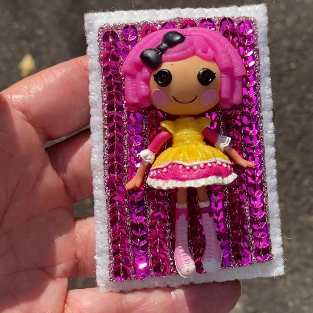 Hand holding a Lalaloopsy doll brooch covered in pink sequins shown in sunlight.