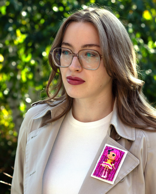 Handmade Lalaloopsy doll brooch covered in pink sequins worn on a trench coat lapel by a young woman.