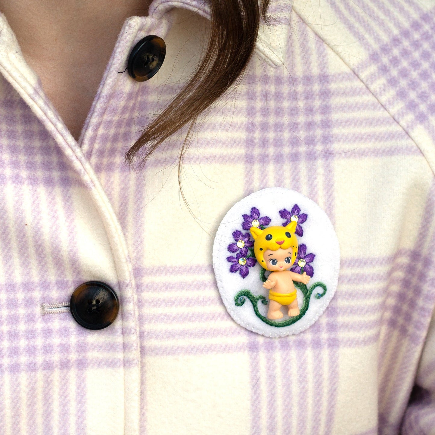Closeup of a handmade embroidered floral brooch featuring Lippy Twozie worn on a plaid coat.