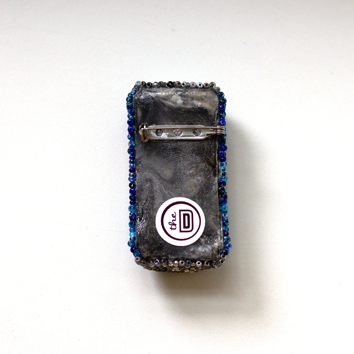 The back of a beaded Pepsi can brooch with a metal clasp, shown on a plain white background. A circular white sticker with 'The DD' brand is attached near the bottom.