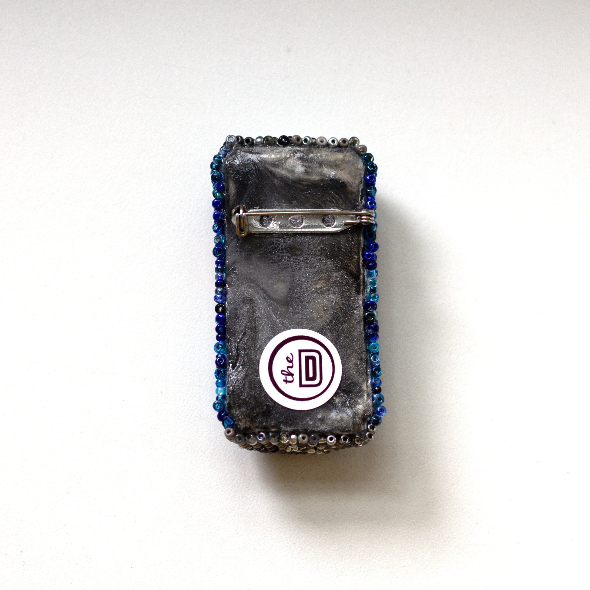 The back of a beaded Pepsi can brooch with a metal clasp, shown on a plain white background. A circular white sticker with 'The DD' brand is attached near the bottom.