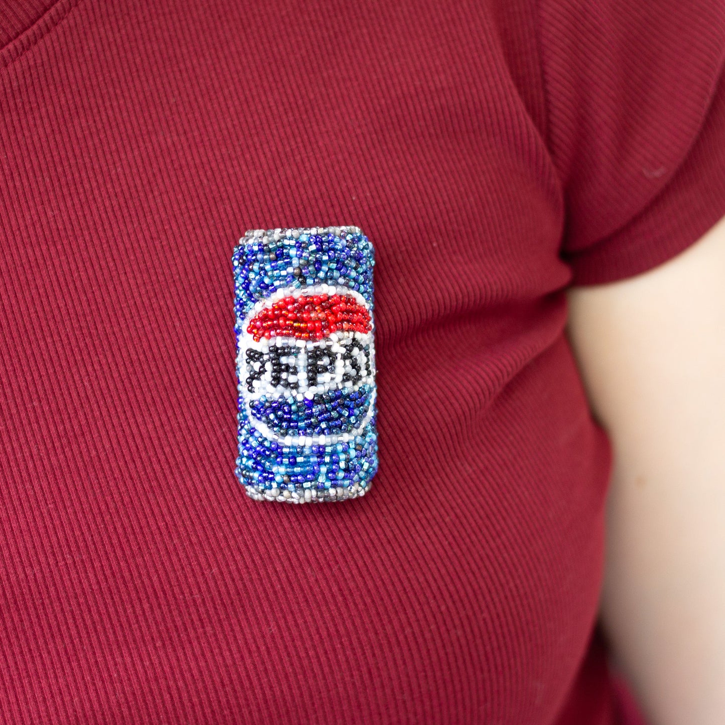 A close-up of a person wearing a red t-shirt with a small, beaded Pepsi can pin on the chest.