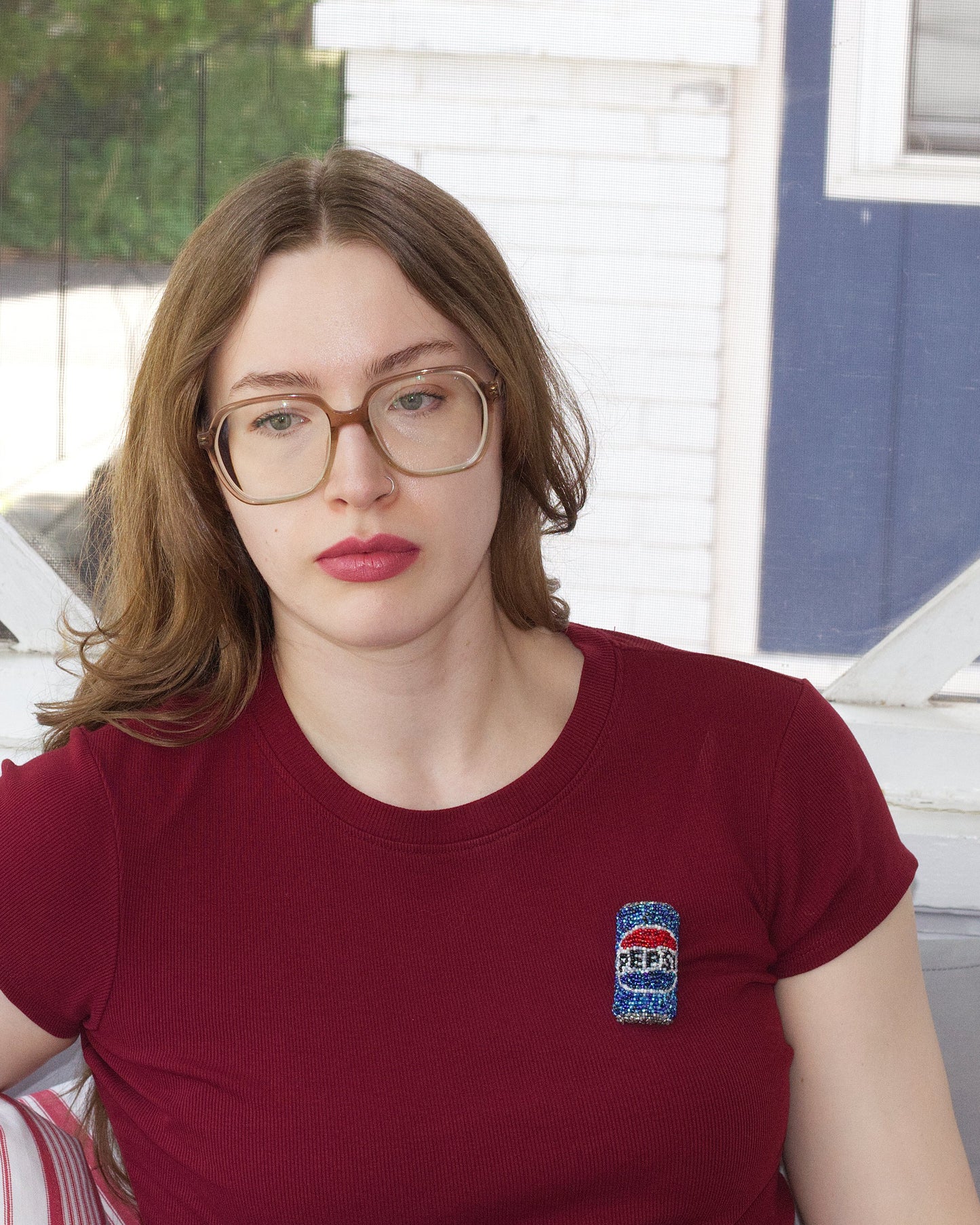 A woman with glasses and brown hair sits indoors wearing a burgundy t-shirt with a beaded Pepsi can brooch on the left chest. She is positioned in front of white-framed windows with a blue and white exterior wall visible in the background.