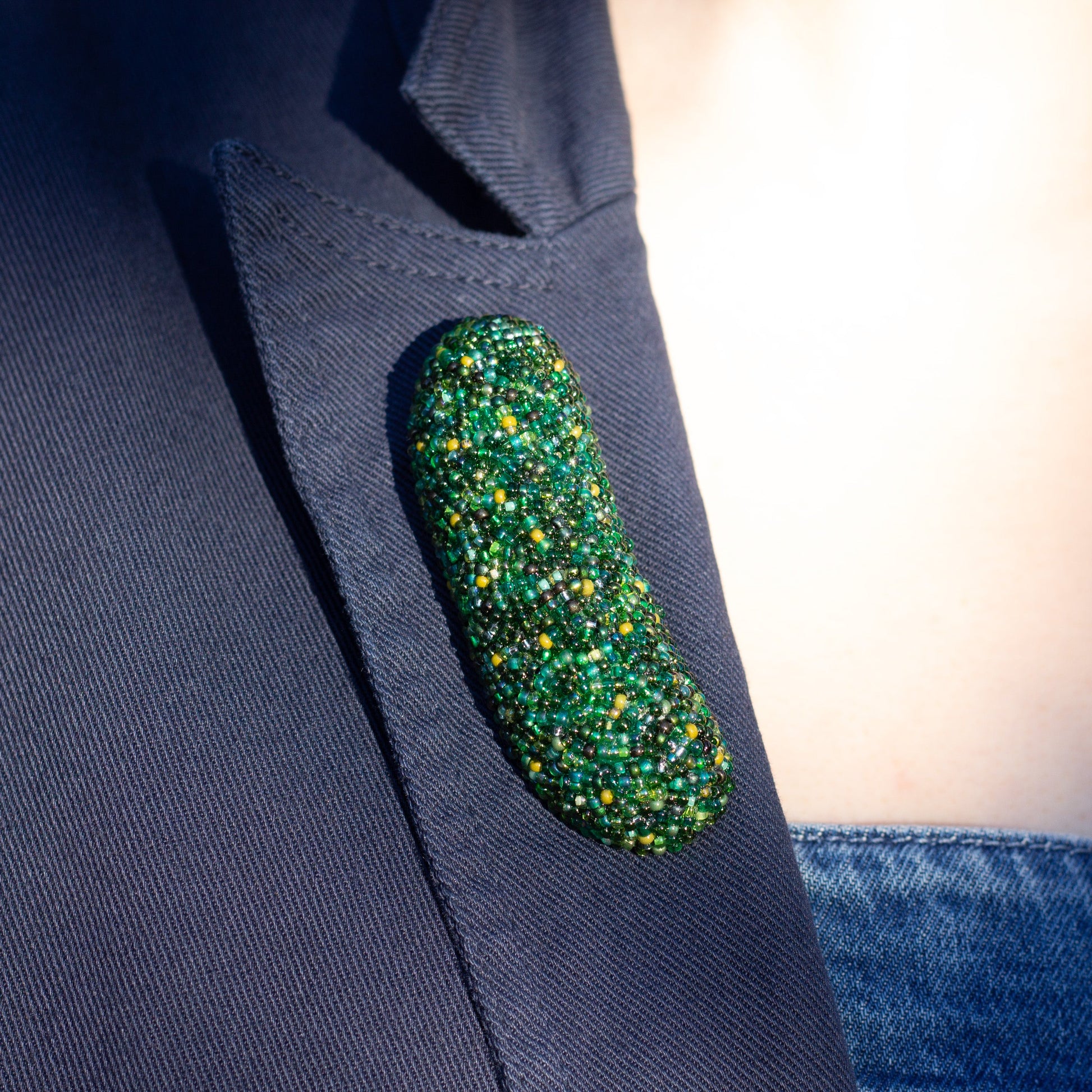 Closeup of a handmade beaded pickle brooch worn on the lapel of a blue blazer.