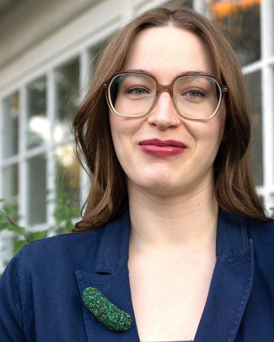 Handmade beaded pickle brooch worn on the lapel of a blue blazer by a young woman.