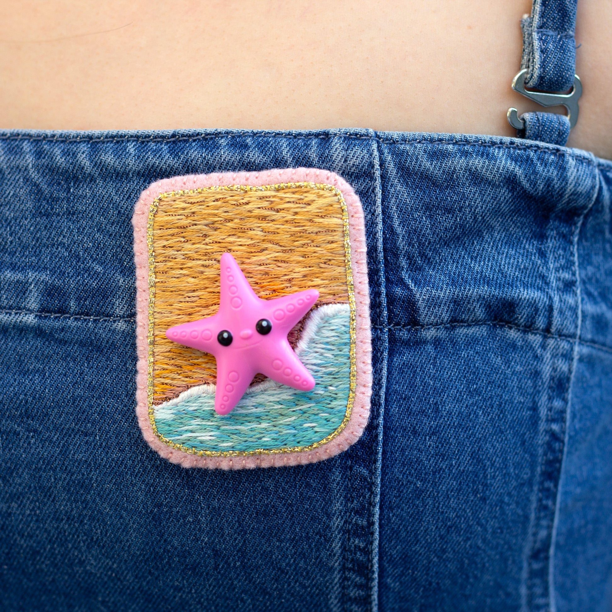 Closeup view of a handmade embroidered beach scene brooch featuring a starfish worn on a denim dress.