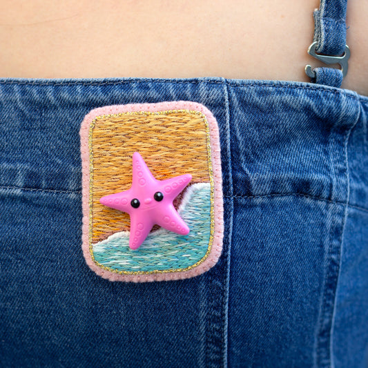 Closeup view of a handmade embroidered beach scene brooch featuring a starfish worn on a denim dress.