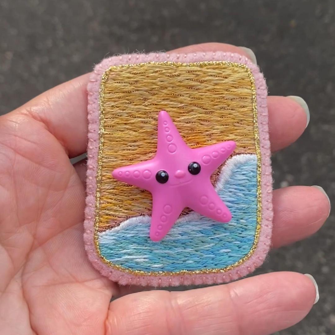 Hand holding an embroidered beach scene brooch featuring a starfish shown in sunlight.